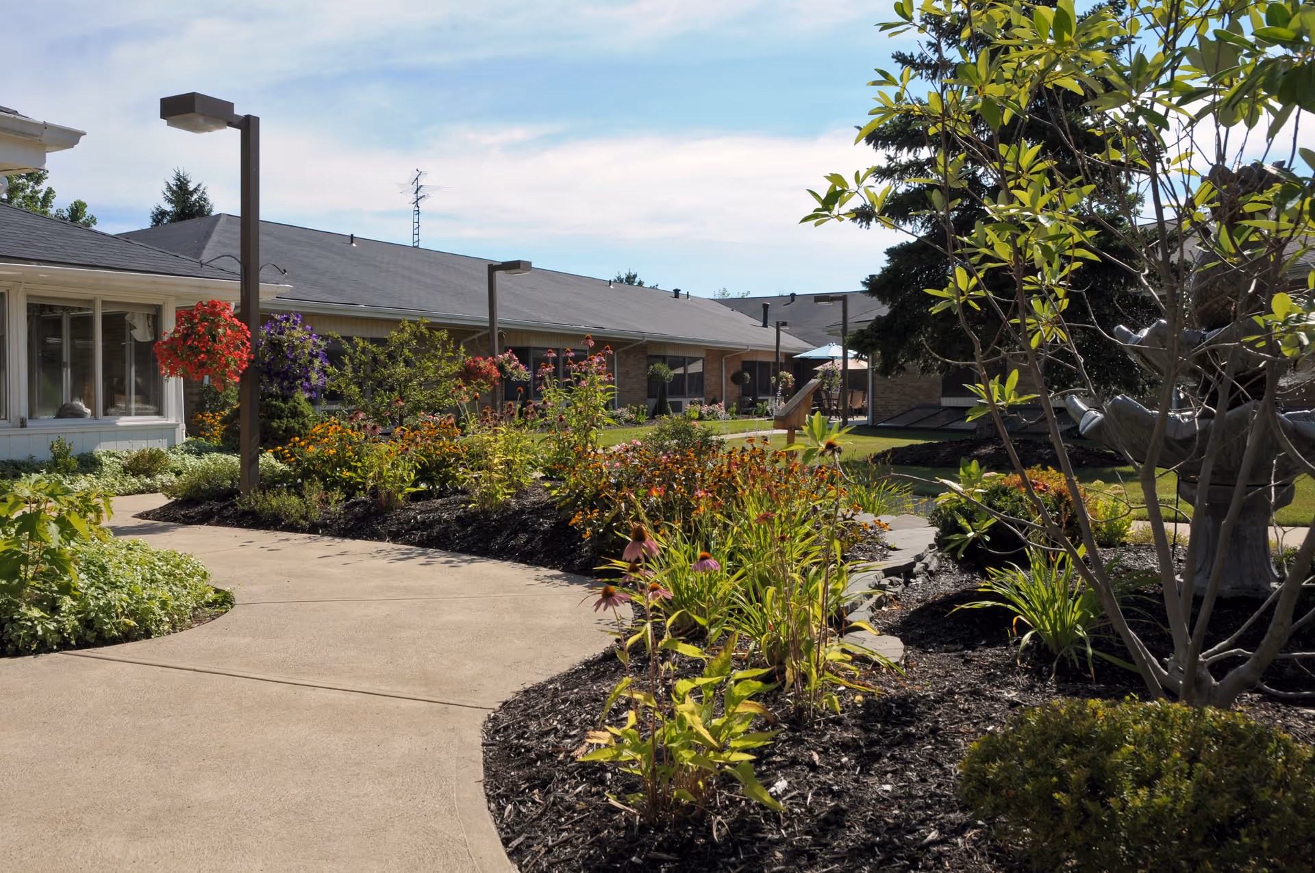 Sunlit landscaped courtyard with winding concrete paths, flowerbeds, hanging baskets and single-story facility buildings in the background.