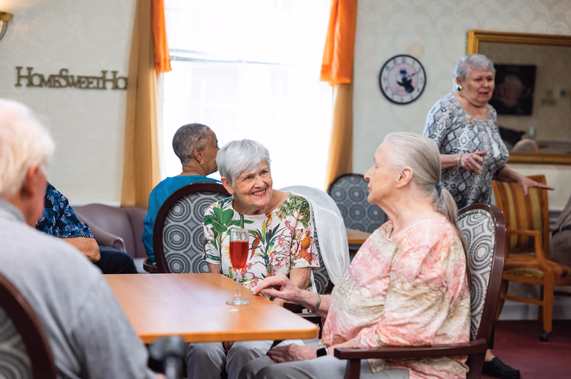 A group of elderly people sitting and chatting in a cozy common room with patterned chairs and a wooden table. One woman is smiling and holding a glass with a red drink. The room has warm lighting, orange curtains, a wall clock, and a decorative sign that reads 'Home Sweet Home'.