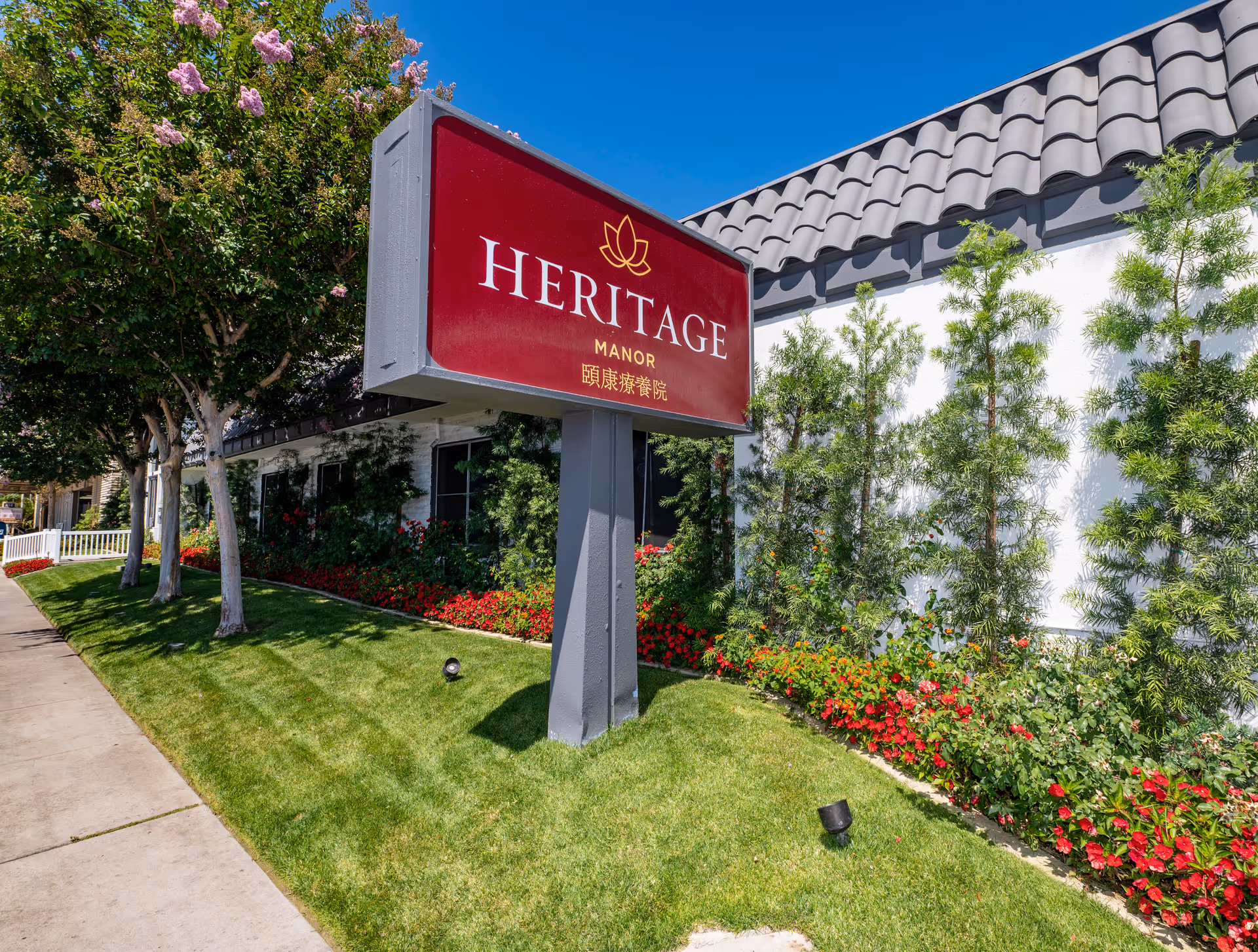 Outdoor view of Heritage Manor 頤康療養院 facility sign on a green lawn with trees, flowers, and a building with a tiled roof in the background under a clear blue sky.