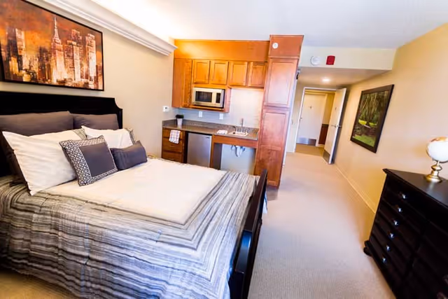 A cozy studio apartment featuring a neatly made bed with multiple pillows and a striped comforter. Behind the bed is a large framed cityscape artwork. To the right of the bed is a compact kitchenette with wooden cabinets, a microwave, a small refrigerator, and a sink. The room has beige walls and carpeted flooring. A hallway extends from the kitchenette area, with a framed nature painting on the wall and a dark wooden dresser with a globe on top.