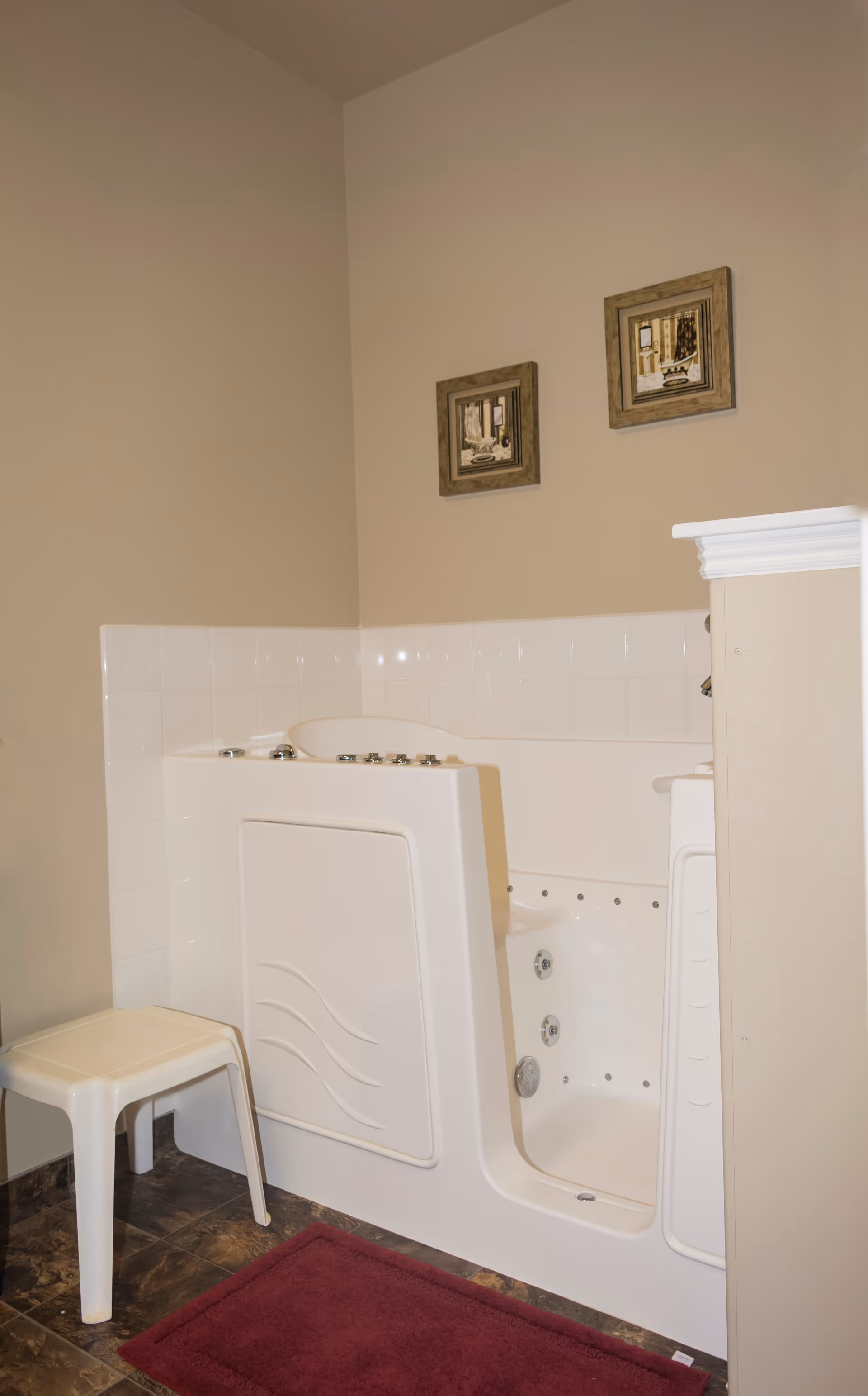 A walk-in bathtub with jets and a door for easy access is installed in a bathroom corner. There is a small white plastic stool next to the tub, a burgundy bath mat on the floor, and two framed pictures hanging on the beige wall above the tub.