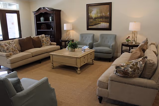 A cozy senior living facility common area with two beige sofas adorned with patterned and solid pillows, two light blue armchairs, a wooden coffee table with a plant and books, two side tables each with a lamp, a dark wooden bookshelf, and a framed landscape painting on the wall.