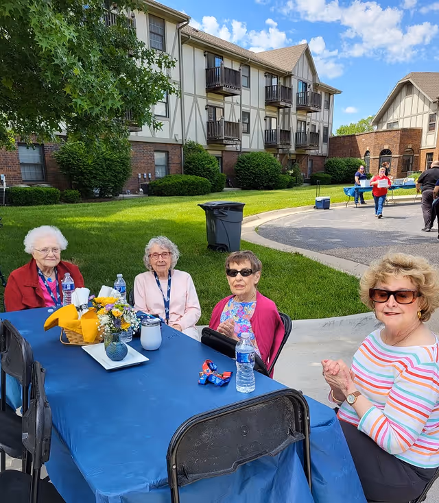 Four elderly women sitting around a blue tablecloth-covered table outdoors near a residential building with Tudor-style architecture. The table has a small flower arrangement, water bottles, and napkins. Other people and tables are visible in the background on a sunny day with a blue sky and scattered clouds.