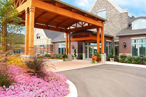Entrance of a senior living facility named Waterford Place featuring a large wooden canopy over the driveway, stone and brick exterior walls, large windows, and landscaped flower beds with pink flowers and ornamental grasses.