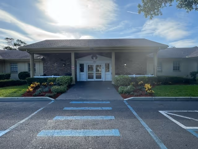 Front exterior entrance of a single-story health and rehabilitation building with a covered drive-up, double glass doors, landscaped beds, and a painted pedestrian crosswalk.