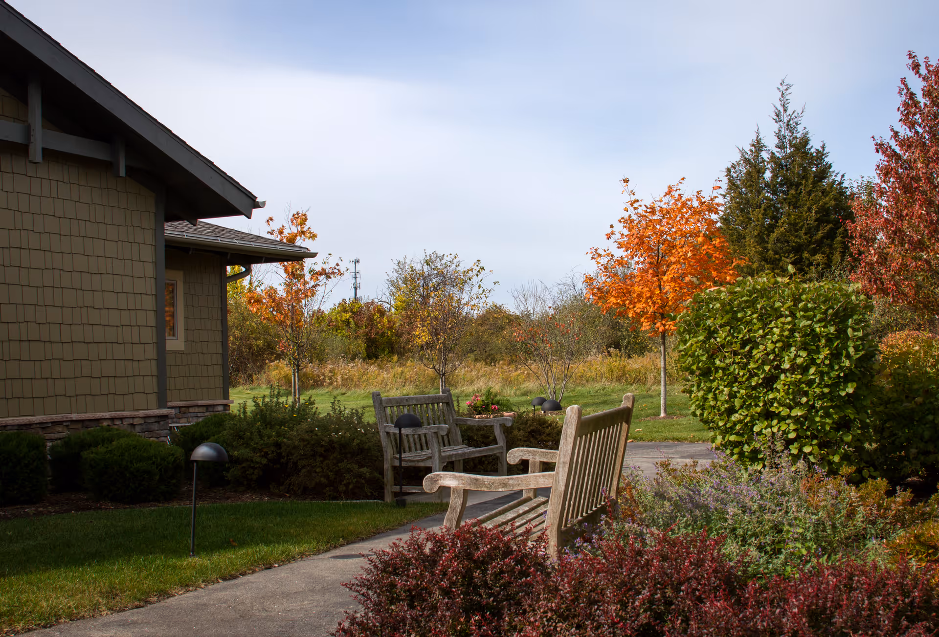 Outdoor seating area with wooden benches along a paved pathway surrounded by green bushes, colorful autumn trees, and a building with brown siding on the left side under a partly cloudy sky.