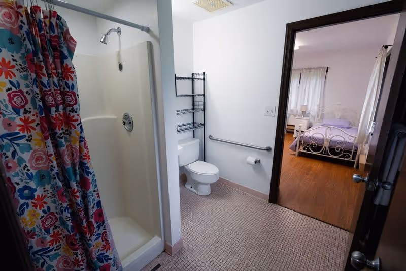 Bathroom with a shower stall and colorful curtain, toilet with grab bar and shelving, and an open door leading to a bedroom.
