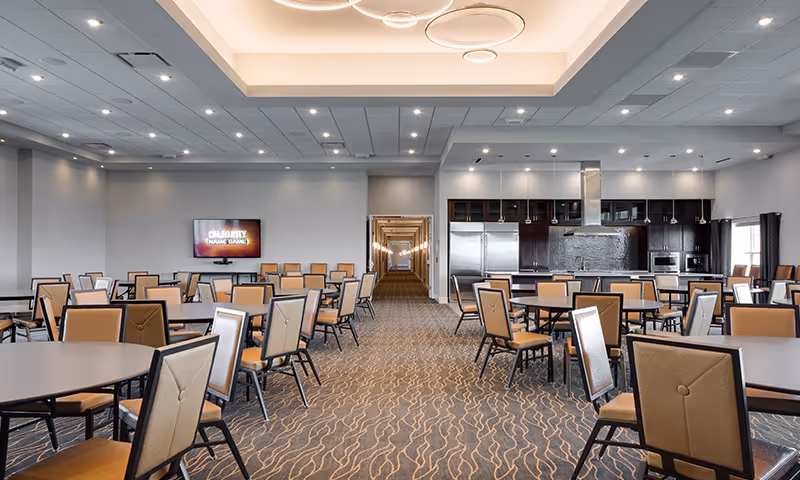 A spacious dining room with multiple round tables and beige cushioned chairs arranged neatly. At the far end, there is a modern kitchen area with stainless steel appliances including a refrigerator, oven, and microwave. A flat-screen TV is mounted on the left wall displaying a game titled 'Celebrity Name Game'. The room has a patterned carpet, recessed lighting, and a hallway visible in the center background.