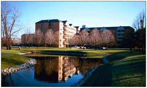 A large multi-story residential building reflected in a calm pond surrounded by green grass and leafless trees under a clear blue sky.