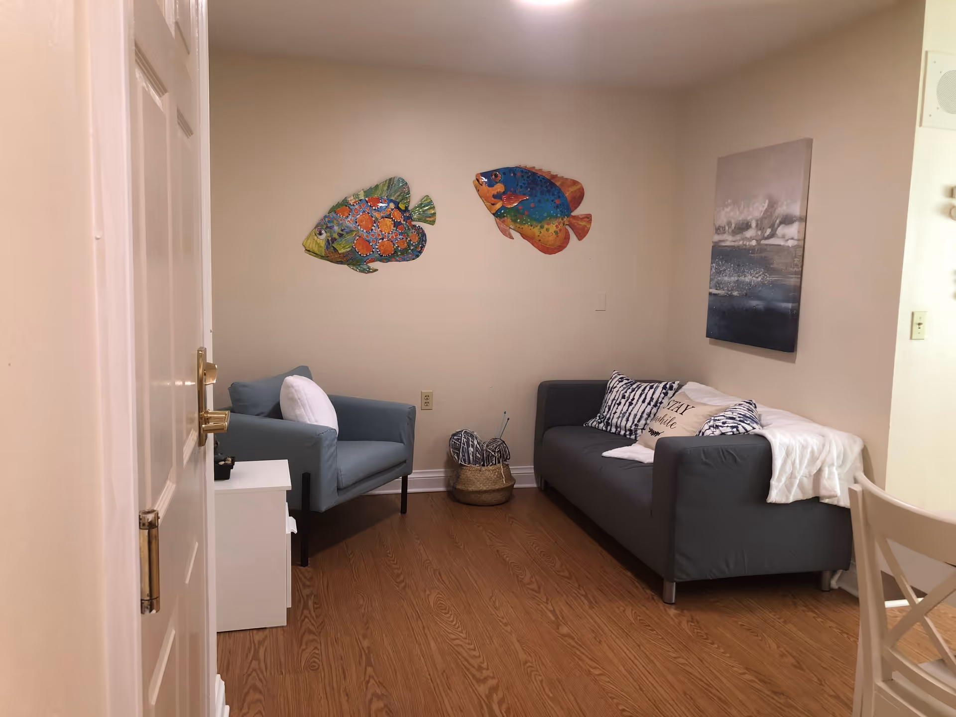 A small living room with a gray couch adorned with patterned and plain pillows, a light blue armchair with a white pillow, and a white side table. The walls are decorated with two colorful fish wall hangings and a framed abstract painting. The floor is wooden, and there is a basket with yarn and knitting needles between the couch and armchair.