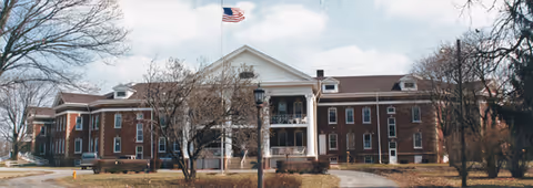 Brick senior living facility with central entrance, columns, and American flag.