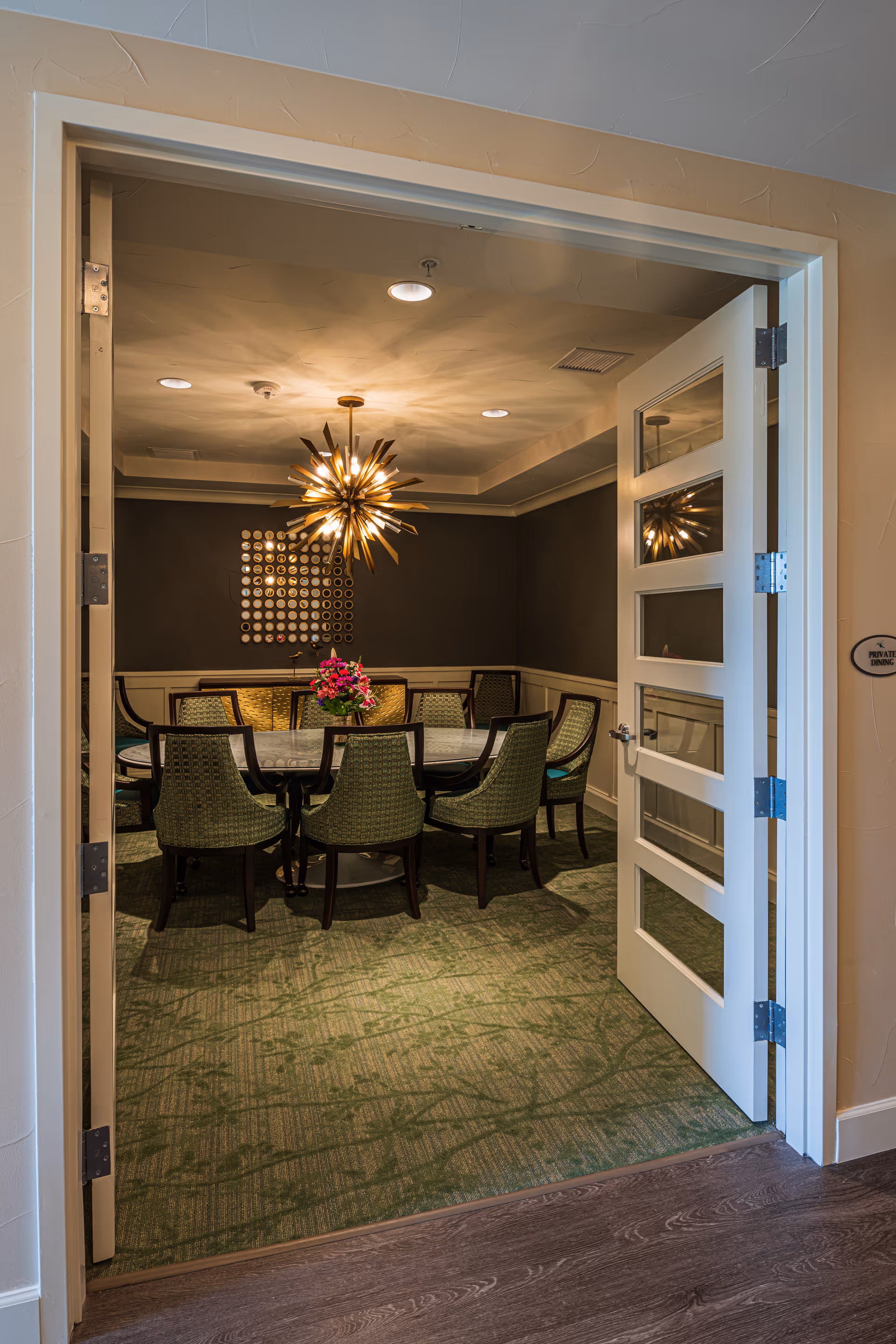 Open double doors reveal a private dining room with a round table surrounded by green upholstered chairs, a starburst chandelier, and a floral centerpiece.