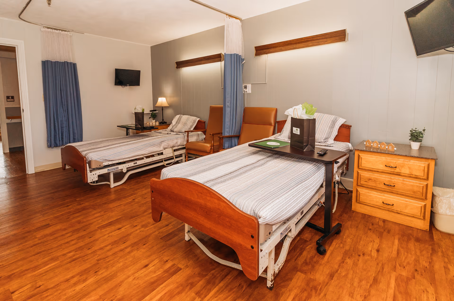 Two hospital-style beds with bedside tables, chairs, and medical curtains in a nursing-home patient room.