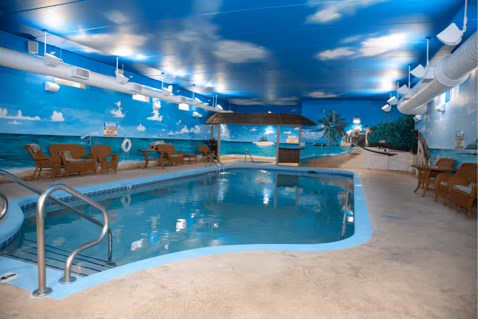 Indoor swimming pool area with a beach-themed mural on the walls and ceiling, featuring ocean waves, palm trees, and a sandy shore. The pool has metal handrails and steps, surrounded by wicker chairs and tables. The ceiling is painted blue with white clouds, and there are ventilation ducts and lights along the walls.