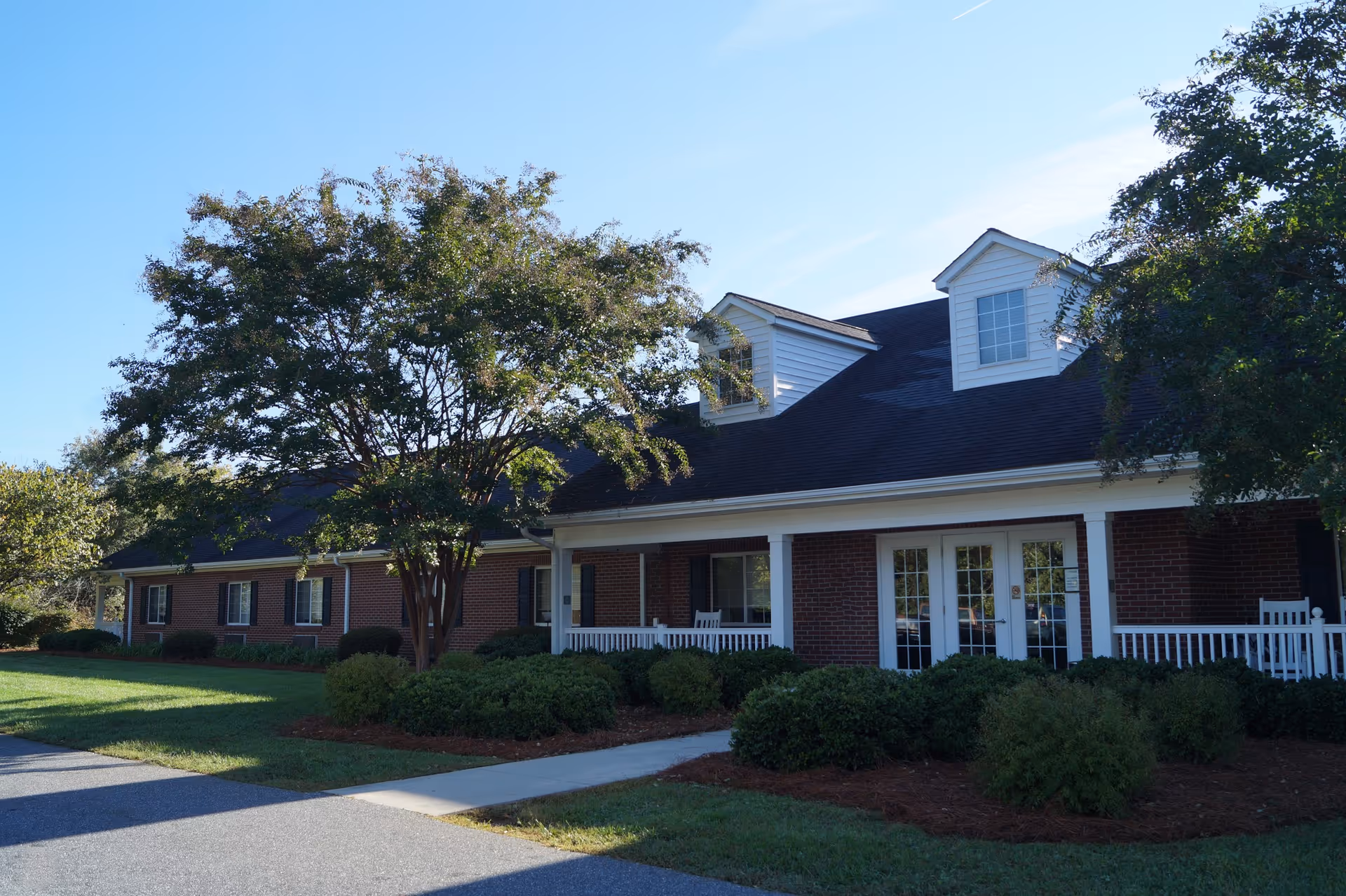Front exterior of a one-story brick building with a covered porch, dormer windows, a walkway, and landscaped shrubs and trees.
