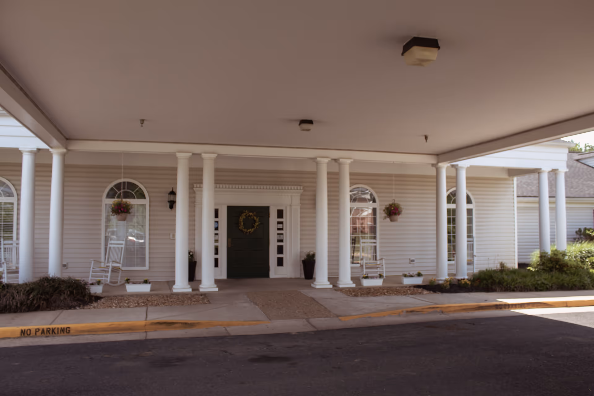Covered entrance of a building with white columns, a dark green door decorated with a wreath, hanging flower baskets, white rocking chairs, and windows with arched tops.