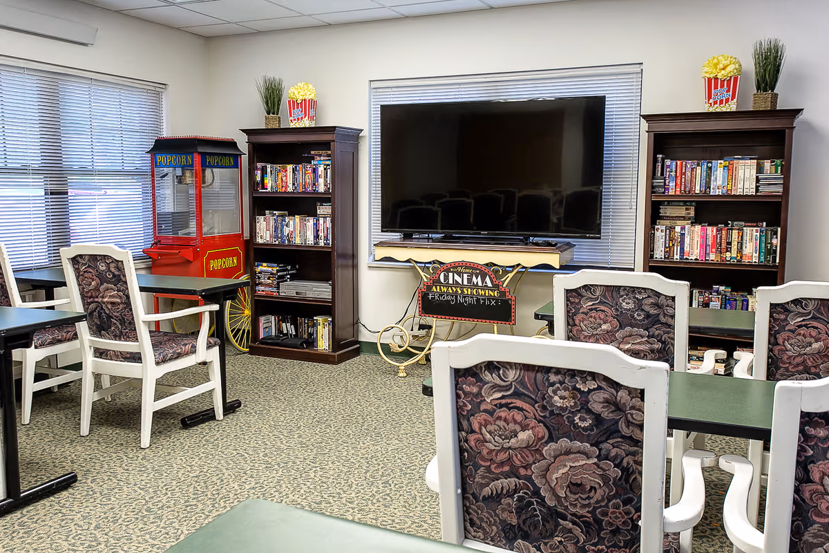 A cozy common room with floral upholstered chairs and green tables facing a large flat-screen TV. There are two wooden bookshelves filled with DVDs and VHS tapes, a vintage-style popcorn machine, and a small sign that reads 'Cinema Always Showing Friday Night Fix'. The room has carpeted flooring and windows with blinds.