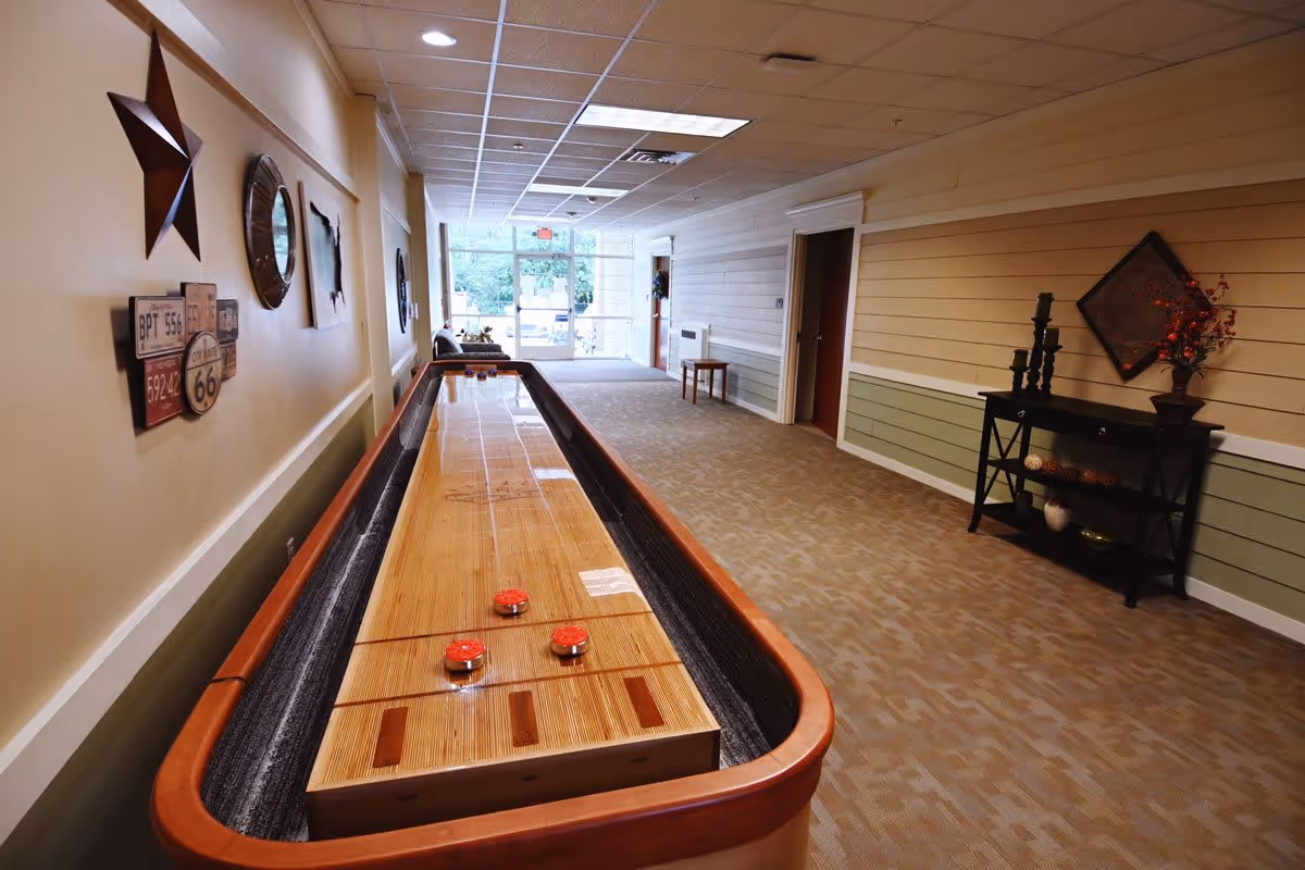 Long indoor common area with a shuffleboard table down the center, wall decor, and seating leading to glass doors at the far end.