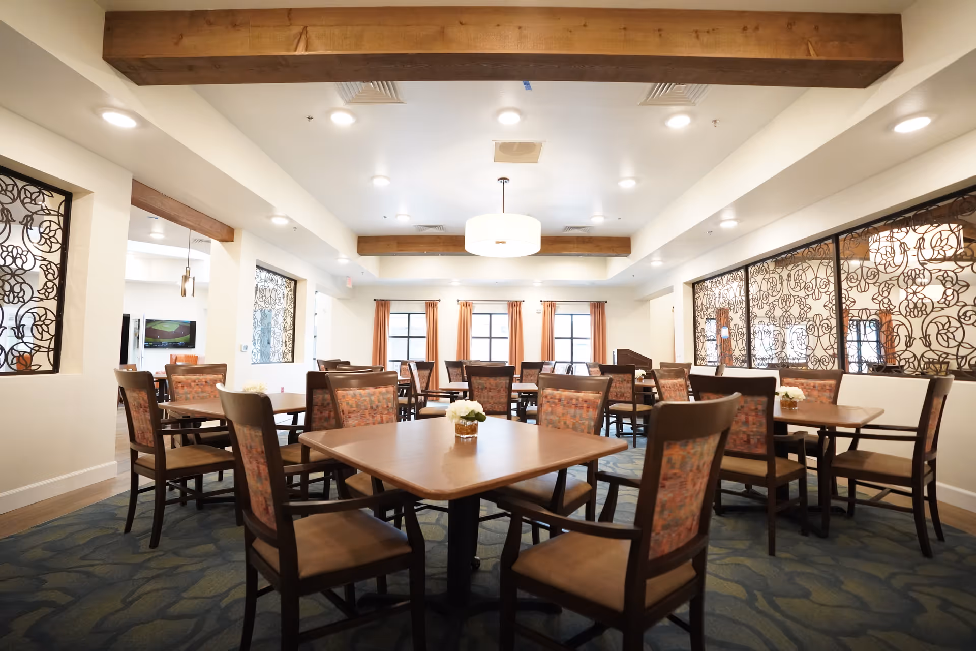A bright dining room with several wooden tables and chairs arranged neatly. The chairs have patterned upholstery, and small flower arrangements are placed on the tables. The room features large windows with orange curtains, decorative wrought iron panels on the walls, exposed wooden ceiling beams, and recessed lighting.
