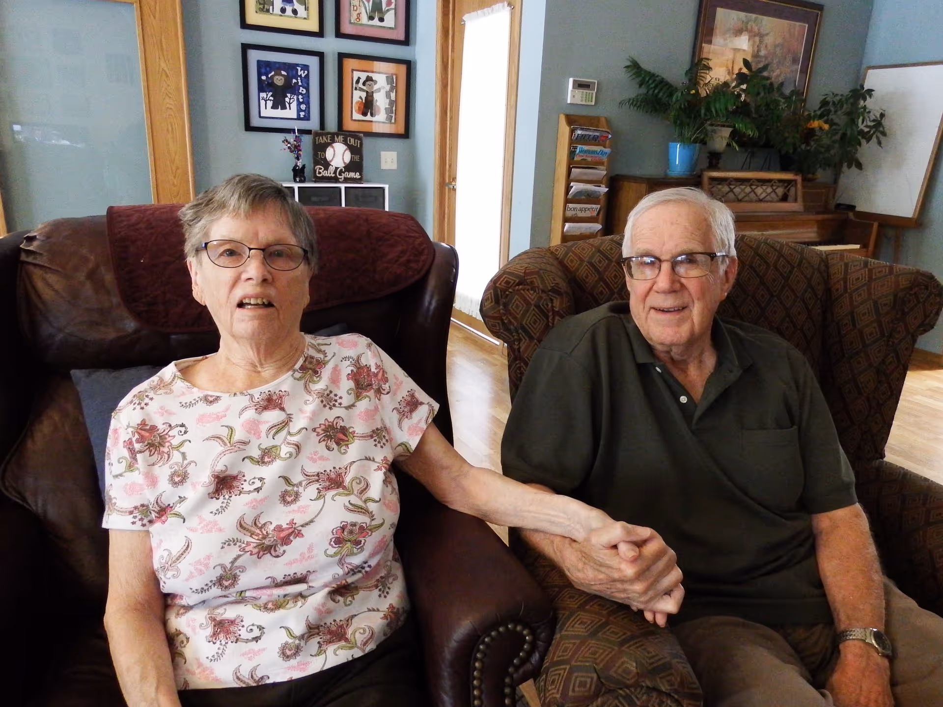 An elderly woman and an elderly man sitting in armchairs in a living room, holding hands. The woman is wearing glasses and a floral patterned shirt, while the man is wearing glasses and a dark green polo shirt. Behind them are framed pictures on the wall, a door with a curtain, a magazine rack, a piano, and some plants.