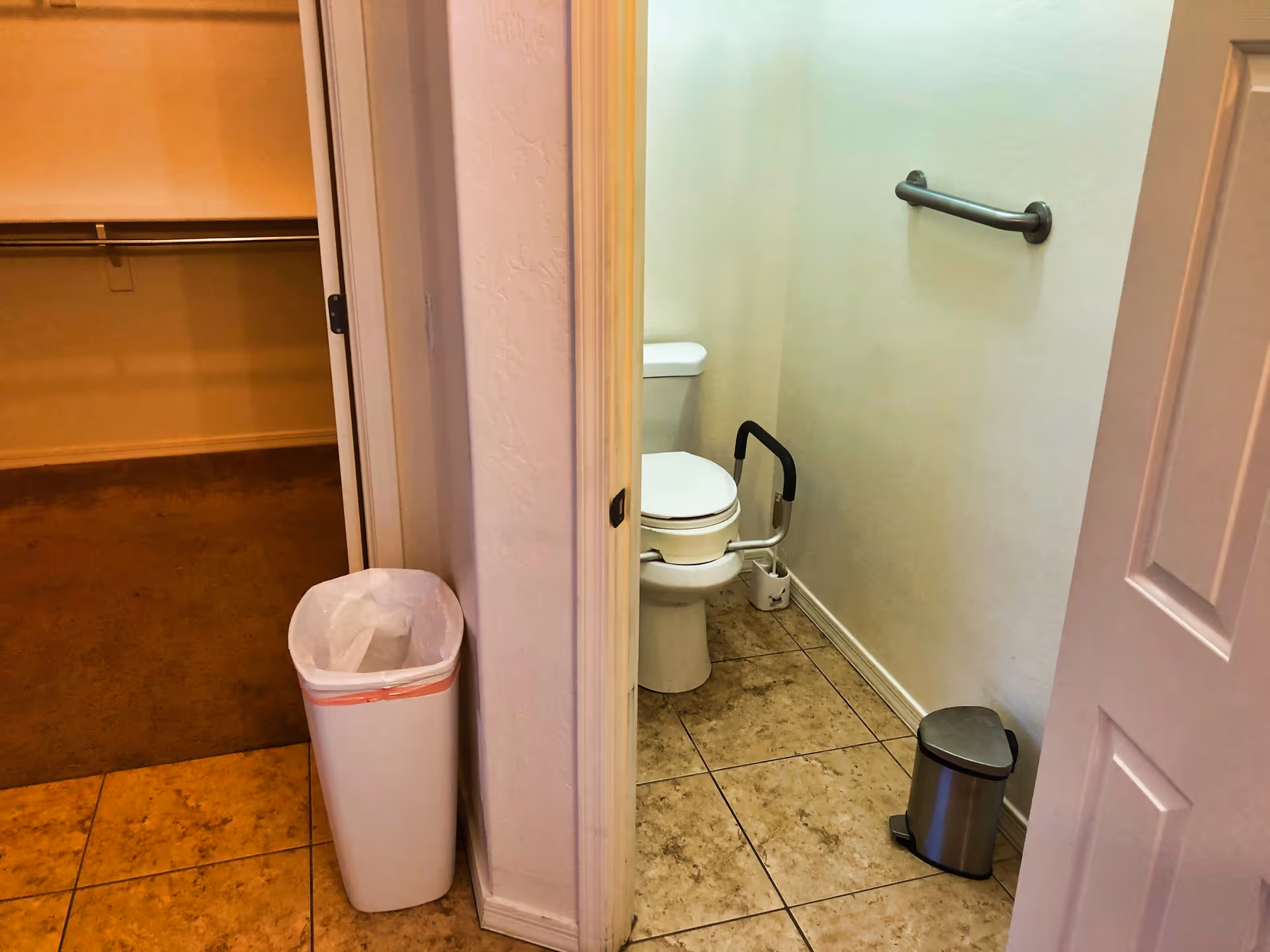 View of a small bathroom with a white toilet equipped with a support rail on the right side, a metal grab bar mounted on the wall, and a small stainless steel trash can on the tiled floor. Adjacent to the bathroom is a closet with carpeted flooring and a white trash bin placed near the doorway.