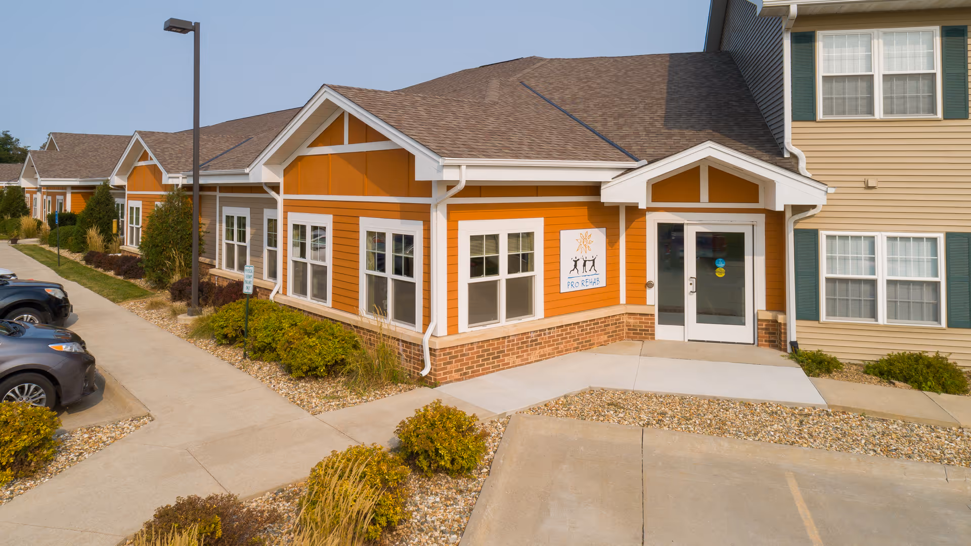 Exterior view of a single-story building with orange siding and white trim, featuring multiple windows and a glass door entrance. There is a parking lot with cars on the left side and landscaped bushes and rocks along the walkway leading to the entrance. The building has a sign that reads 'PRO REHAB' with a logo above it.