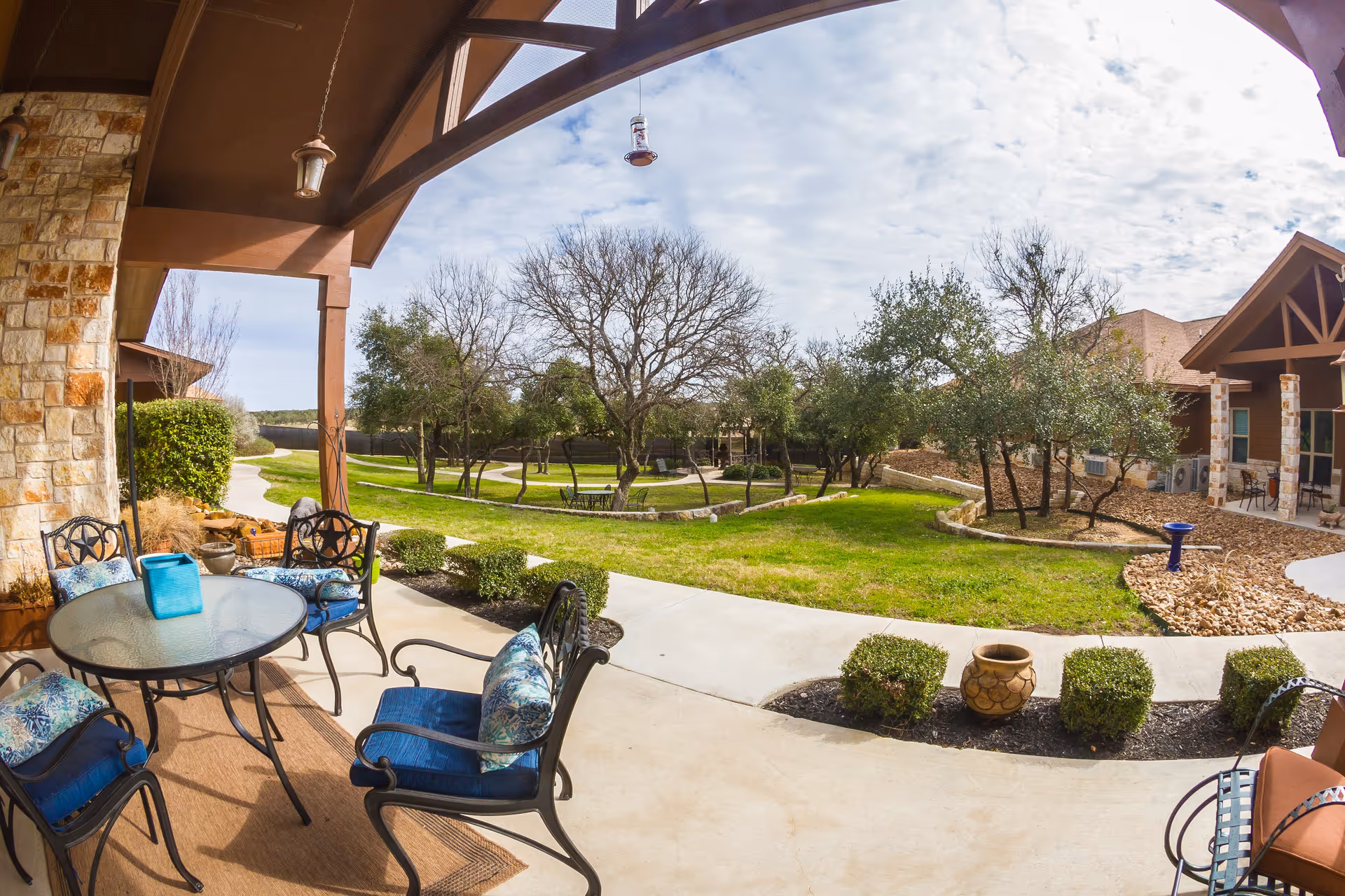 Covered outdoor patio area with a round glass table and four cushioned chairs, overlooking a landscaped garden with trees, shrubs, and a walking path under a partly cloudy sky.