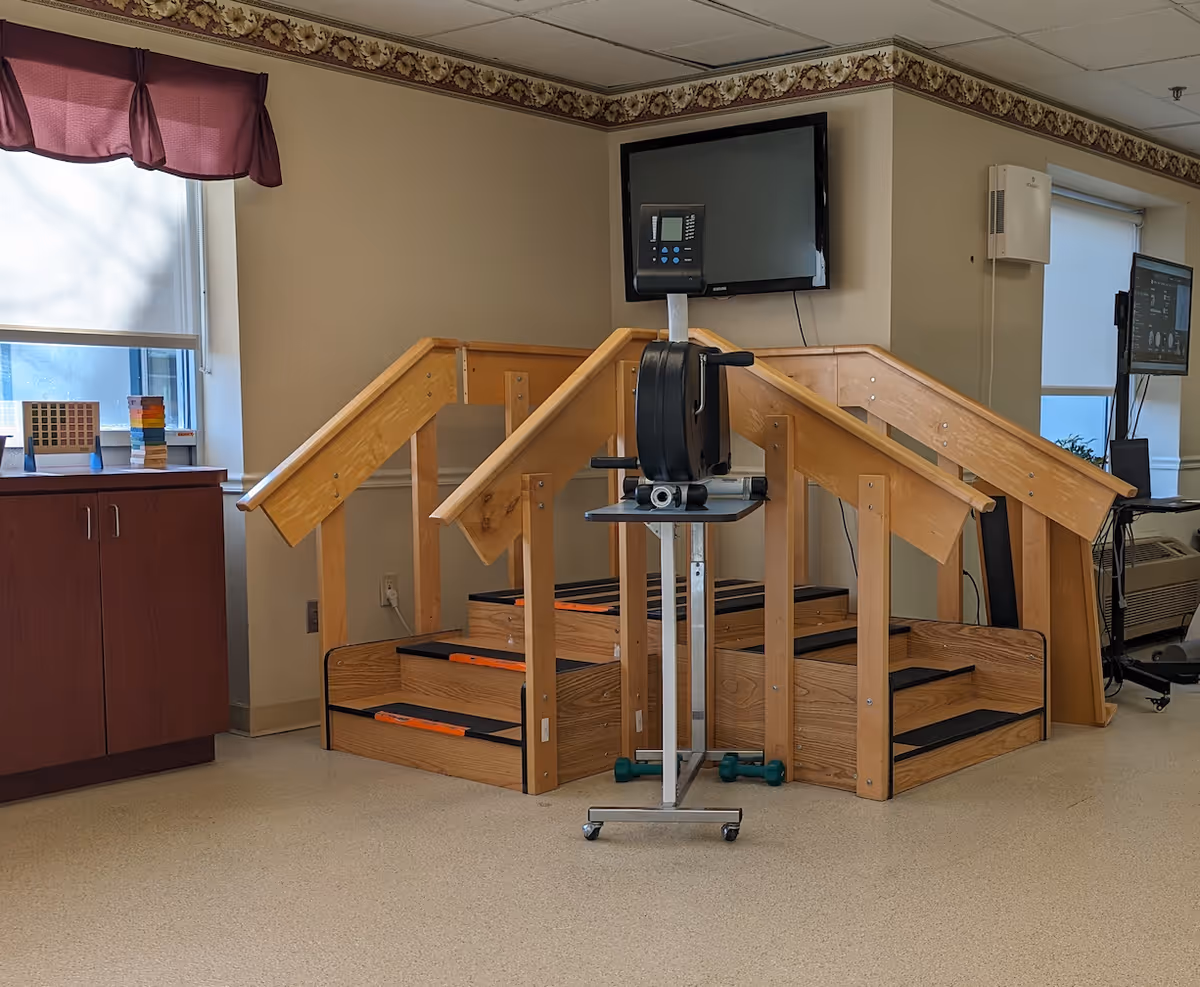 Indoor physical therapy area with wooden stairs and handrails for rehabilitation exercises, a stationary exercise bike on a stand with a monitor, and a cabinet with windows covered by blinds and a maroon valance.