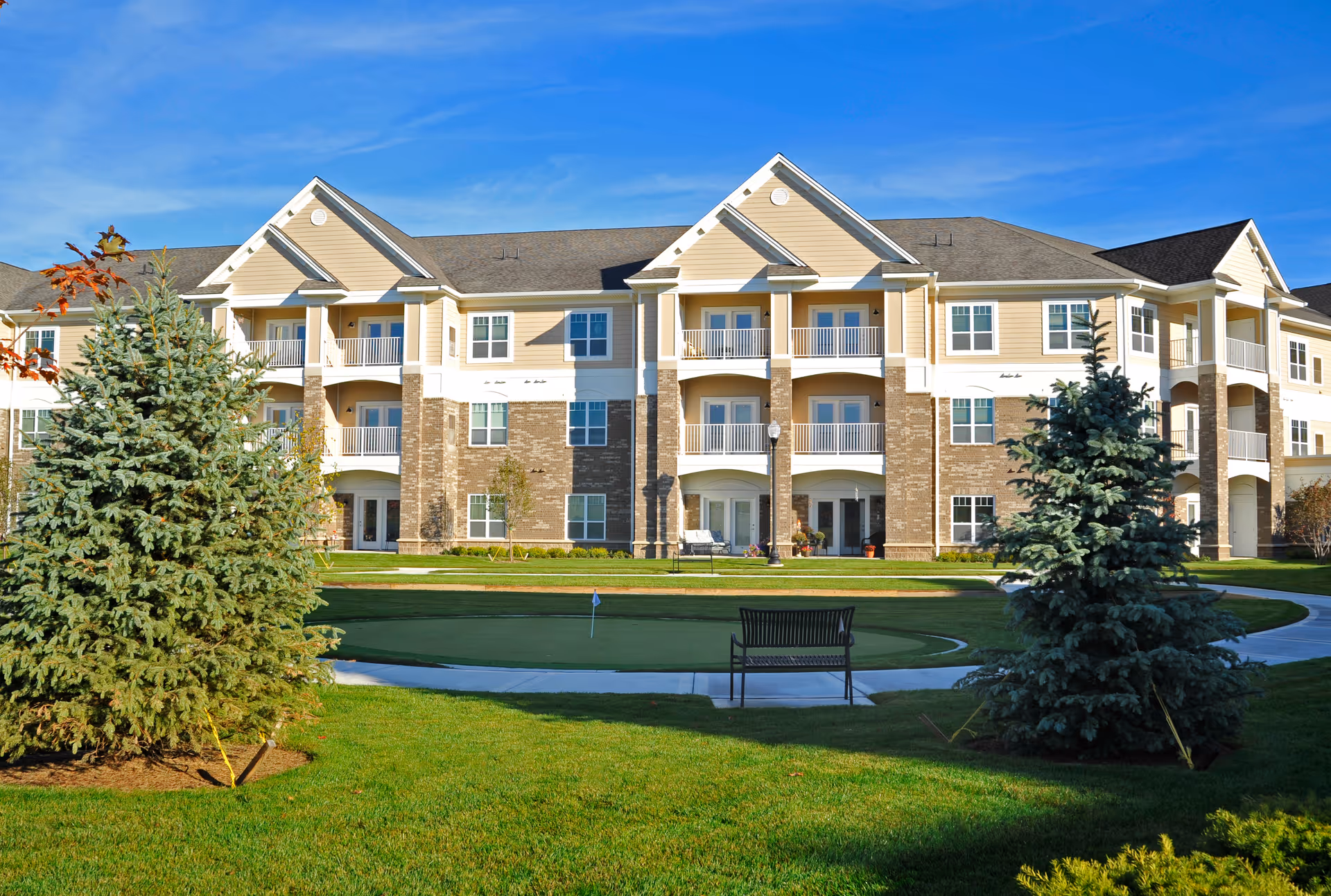 Three-story senior living building with balconies facing a landscaped courtyard with a putting green, bench, and evergreen trees.
