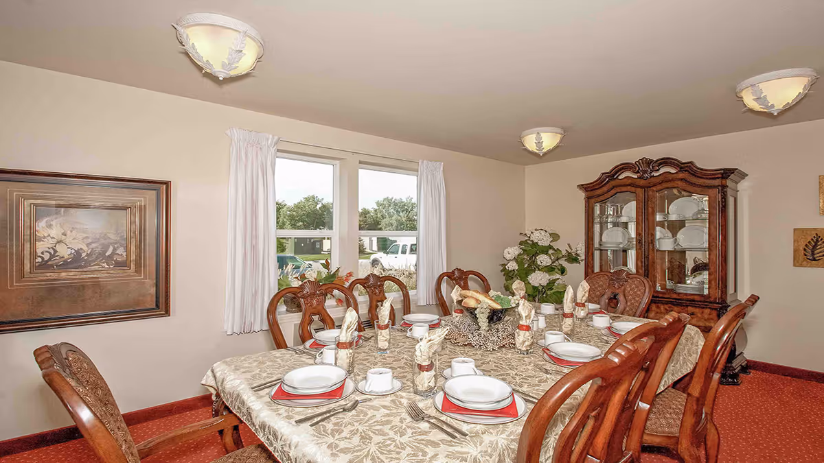 A dining room with a large rectangular table covered with a patterned tablecloth. The table is set with white plates, cups, silverware, and napkins folded in glasses. There are eight wooden chairs around the table. A wooden china cabinet with glass doors displaying dishes is against the wall. Two windows with white curtains let in natural light, and a framed painting hangs on the wall. The room has beige walls, a red carpet, and ceiling lights.