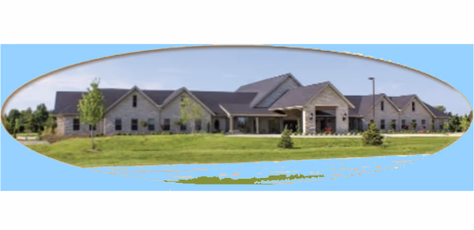 Exterior view of a single-story senior living facility building with a pitched roof, surrounded by a green lawn and small trees under a clear blue sky.