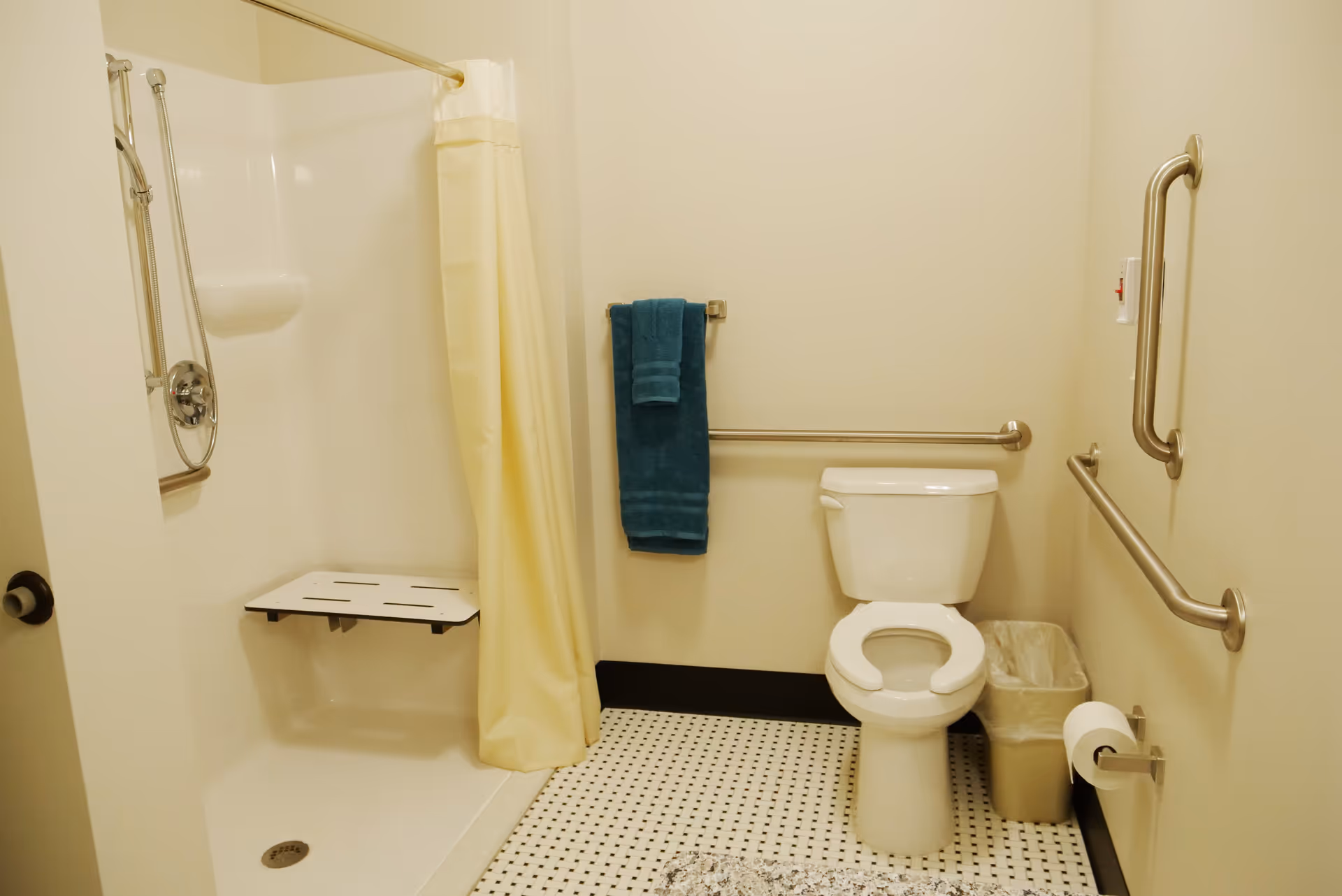 Accessible bathroom with a shower stall and curtain, fold-down shower seat, toilet with grab bars, towel, and waste bin.