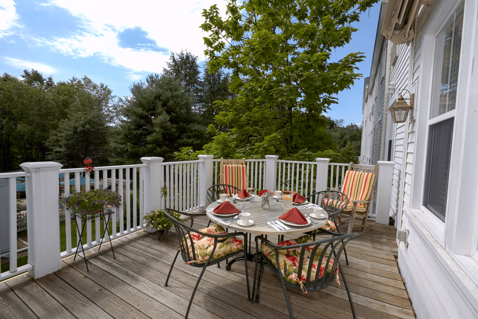 Outdoor wooden deck with a round table set for four with red napkins, cups, and plates. Four chairs with floral and striped cushions surround the table. White railing encloses the deck, and green trees are visible in the background under a partly cloudy sky.