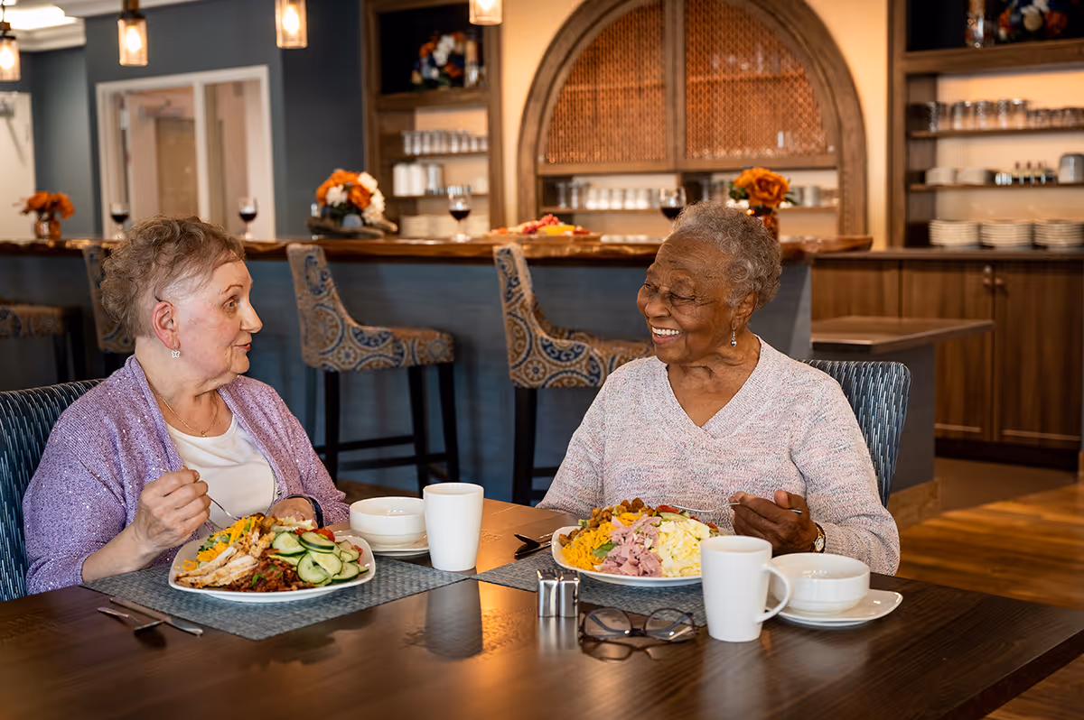 Two elderly women smiling and talking while seated at a dining table with plates of food and coffee cups in a communal dining area.