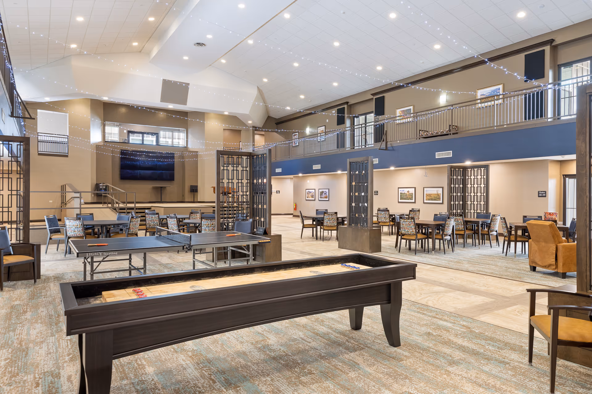 Spacious recreational and dining area in a senior living facility with a shuffleboard table and a ping pong table in the foreground. Several tables and chairs are arranged for dining or socializing, with a large TV mounted on the far wall. The room has high ceilings with string lights hanging across and a second-floor balcony overlooking the space.
