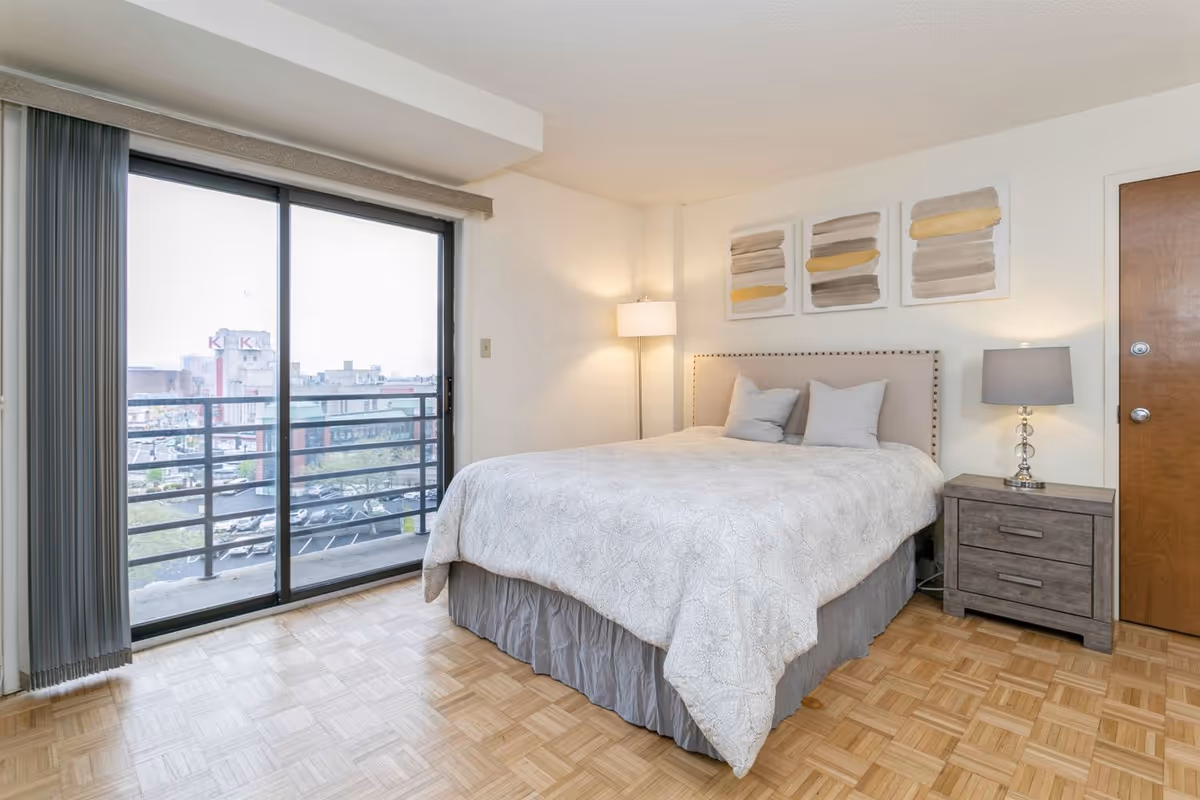 A bright bedroom with a large bed covered in a light-colored quilt and two pillows. There is a wooden nightstand with a lamp on it next to the bed, and a floor lamp in the corner. Three abstract paintings hang above the bed. A sliding glass door with vertical blinds leads to a balcony overlooking a cityscape.
