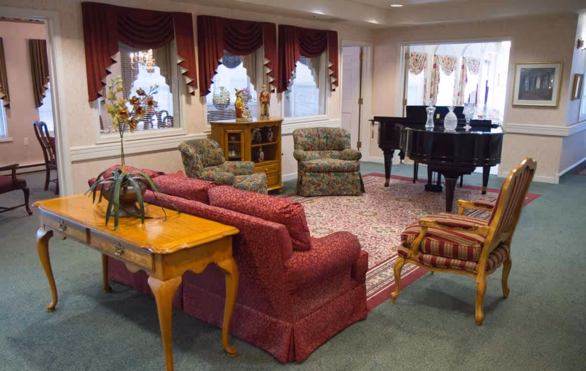 A cozy living room area with a red patterned sofa, two floral armchairs, and a striped wooden chair arranged around a large patterned rug. A black grand piano with glass candle holders is positioned near the back wall. The room has windows with red valances and a wooden side table with a plant on it.