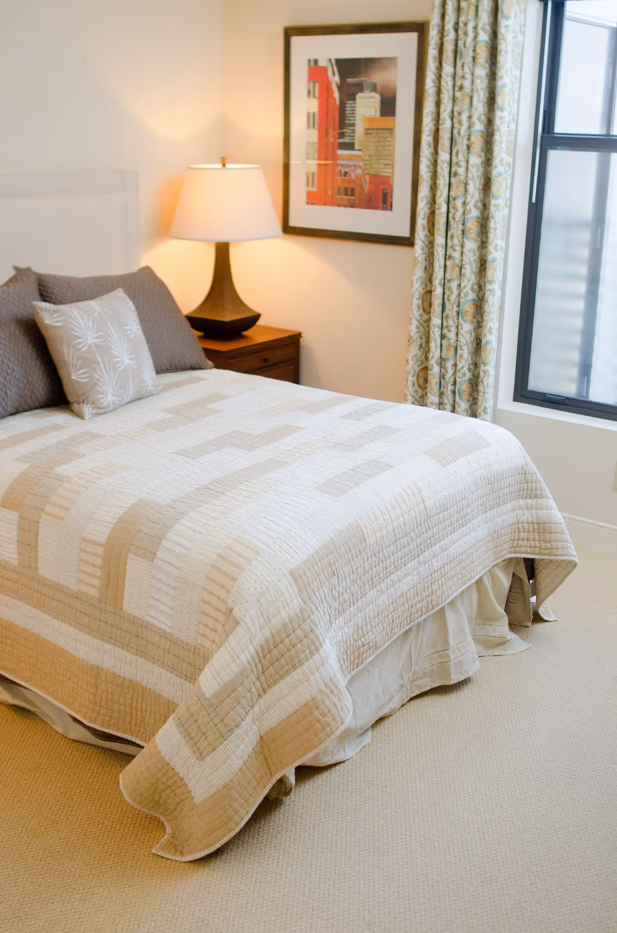 Sunlit bedroom with a neatly made bed, bedside table and lamp, framed artwork, and a window with patterned curtains.