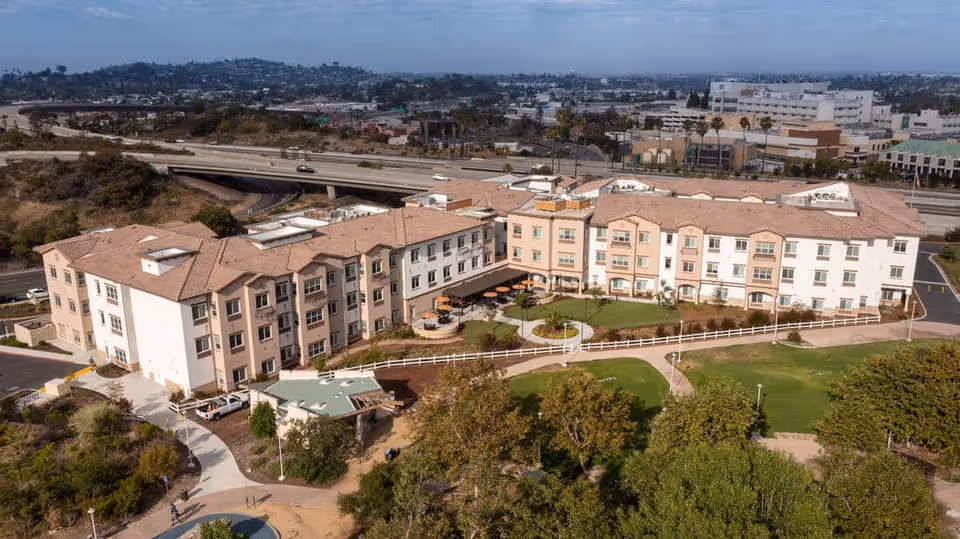 Aerial view of a multi-story senior living building with a central courtyard, landscaped grounds, and a nearby highway.