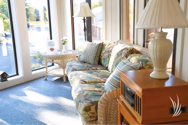 A bright and cozy sitting area with a floral patterned wicker sofa adorned with striped and floral cushions. Next to the sofa is a wicker side table with a small vase of flowers and a lamp. Large windows allow natural light to fill the space, and a wooden cabinet with a lamp is visible in the foreground.