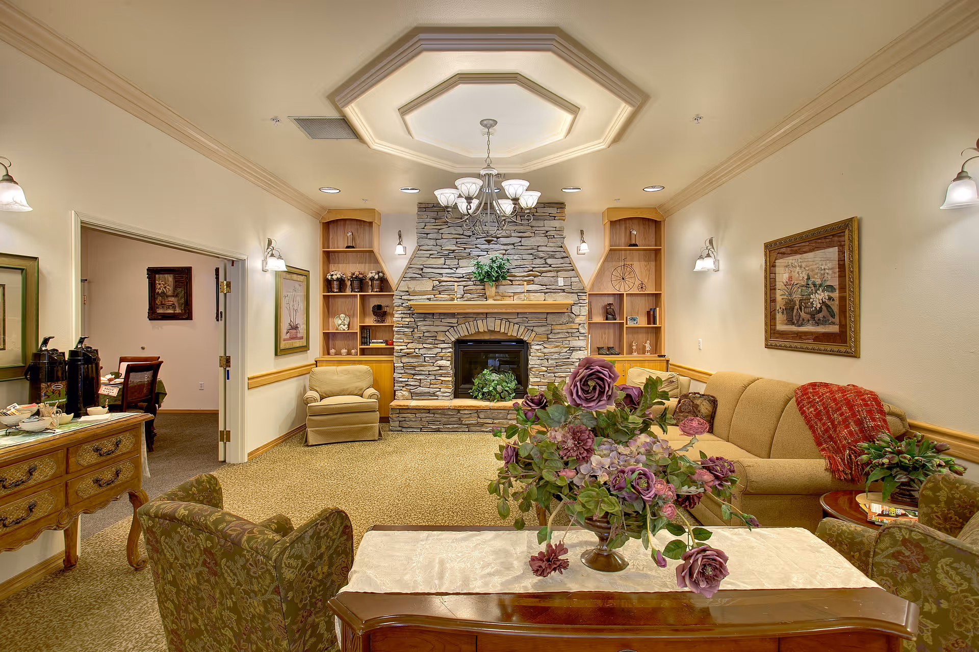 A cozy living room with a stone fireplace centered on the back wall, flanked by built-in wooden shelves. The room features a beige sofa with a red throw blanket, two patterned armchairs, a wooden coffee table with a floral centerpiece, and a chandelier hanging from a decorative ceiling. There are framed pictures on the walls and a wooden sideboard with coffee supplies on the left side.