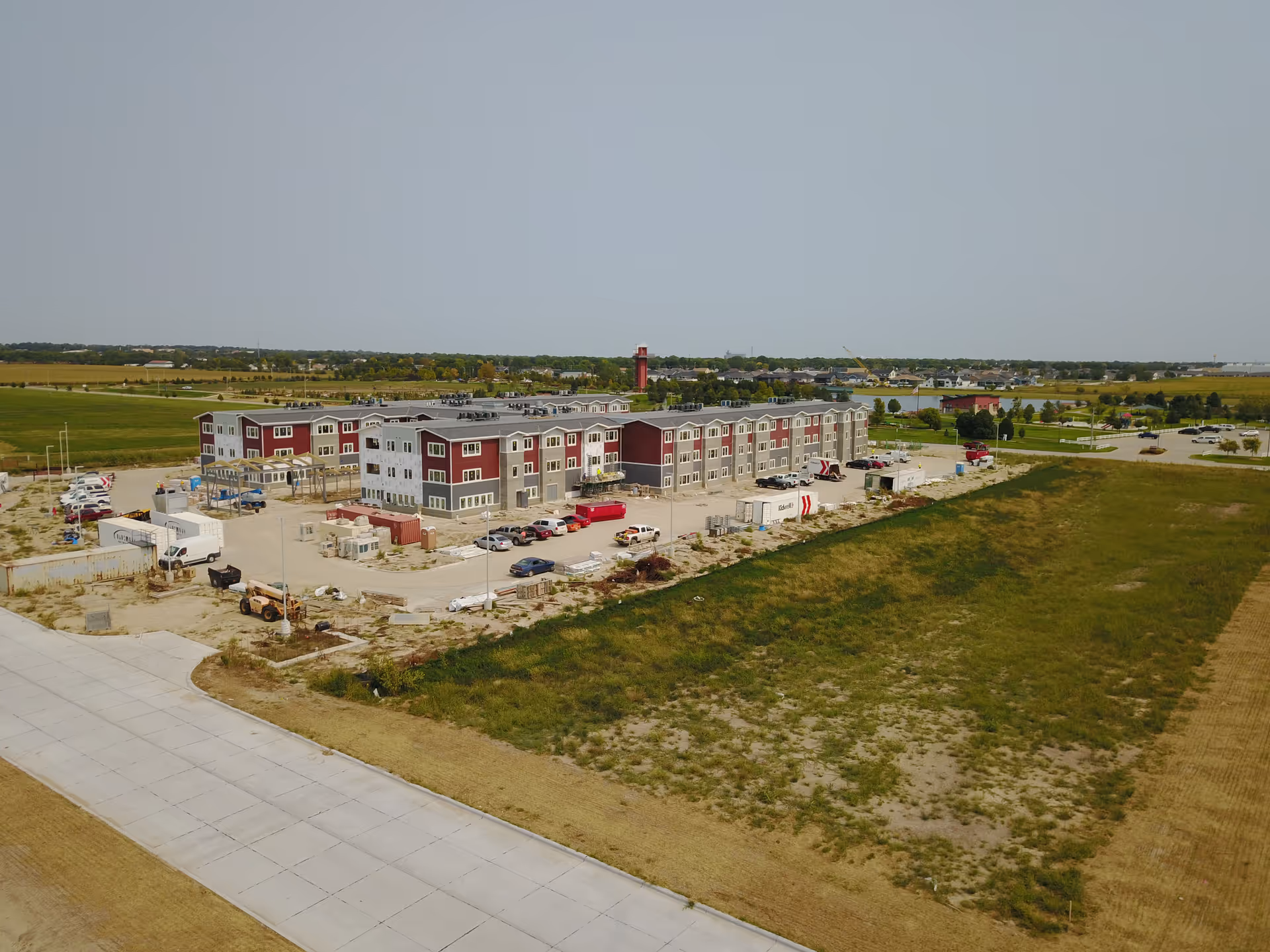 Aerial view of a large multi-story building under construction surrounded by parking areas and open fields, with several vehicles and construction equipment visible around the site.