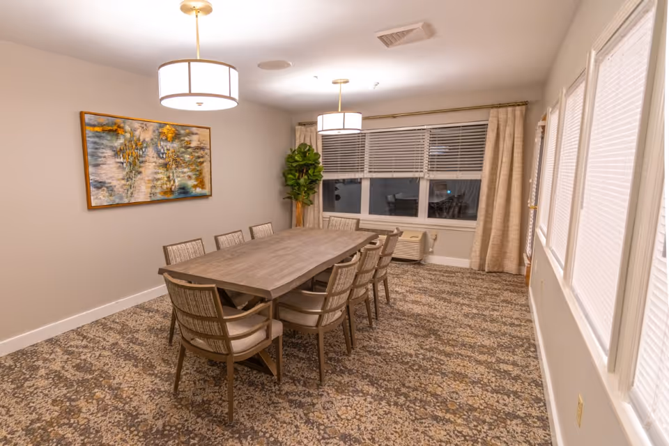 A dining room with a long rectangular wooden table surrounded by eight cushioned chairs. The room has patterned carpet flooring, two modern pendant lights hanging from the ceiling, a large window with blinds and curtains, a potted plant in the corner, and a colorful abstract painting on the wall.