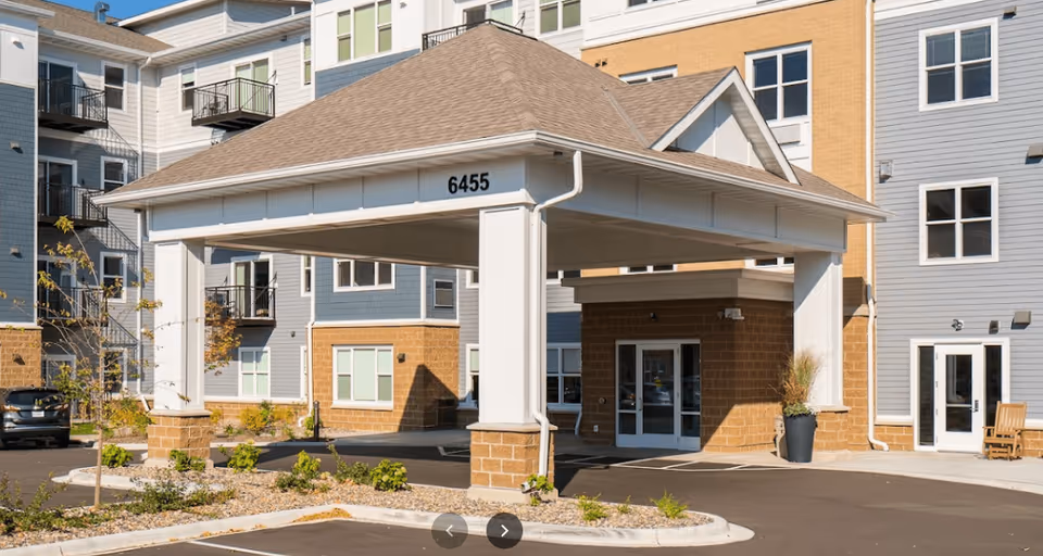 Entrance of a senior living facility with a covered drop-off area supported by white columns and a building number 6455 displayed on the structure. The building has multiple floors with windows and balconies, and there are some plants and a parking area in front.