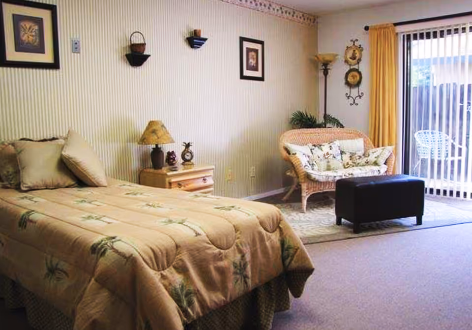 A furnished bedroom with a bed and nightstand, wicker loveseat and ottoman, lamp and wall decor, and a sliding glass door with vertical blinds.