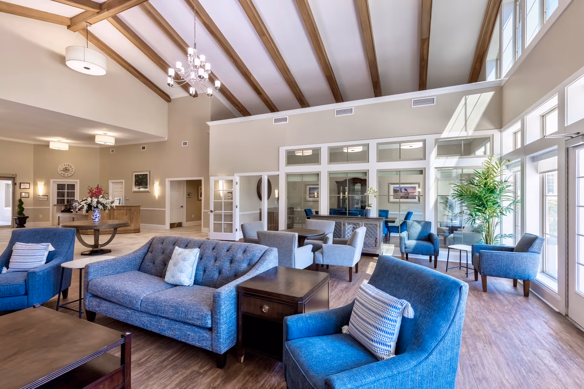 A spacious and well-lit living room area with high vaulted ceilings featuring wooden beams. The room is furnished with blue upholstered sofas and armchairs, wooden coffee tables, and side tables. Large windows on the right side allow natural light to fill the space, and there are plants and decorative items placed around the room. In the background, there is a reception desk and a glass-enclosed room with additional seating.
