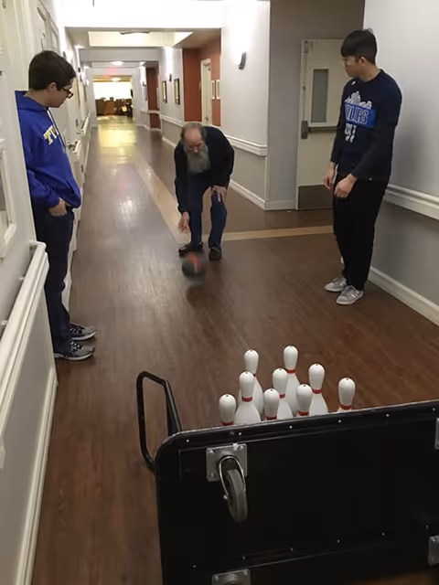 An elderly man is rolling a bowling ball down a hallway towards a set of bowling pins arranged on the floor. Two younger men stand on either side watching him. The hallway has wooden flooring and white walls with handrails.