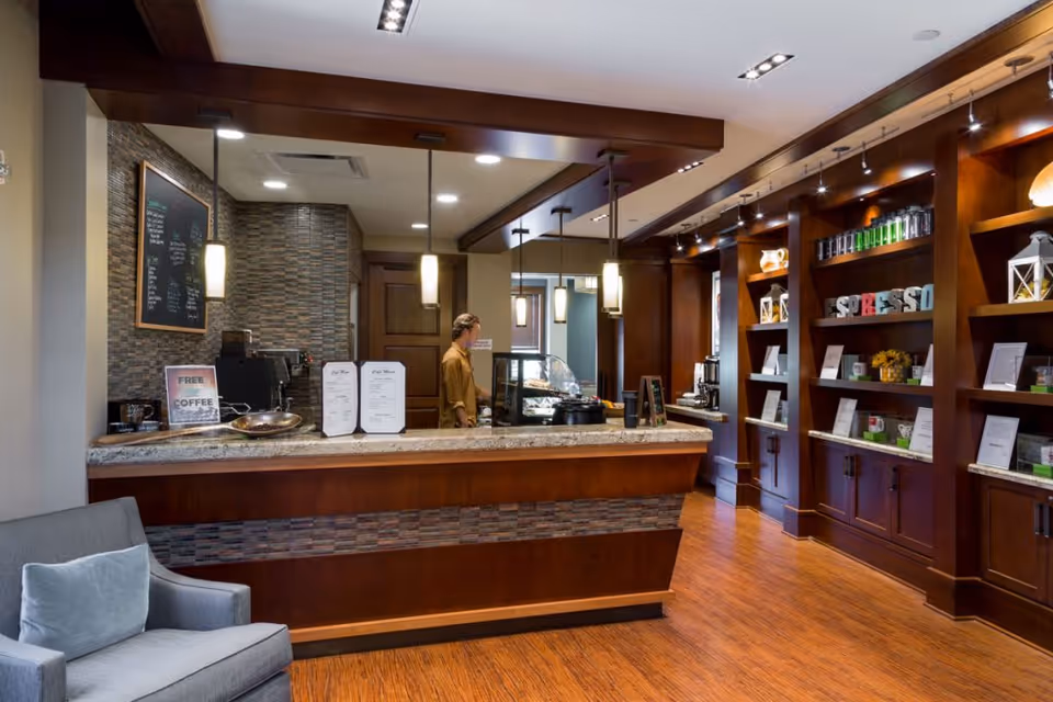Interior view of a coffee bar area in a senior living facility with a wooden counter, a person standing behind the counter, shelves with decorative items and coffee products, pendant lights hanging from the ceiling, and a gray armchair in the foreground.