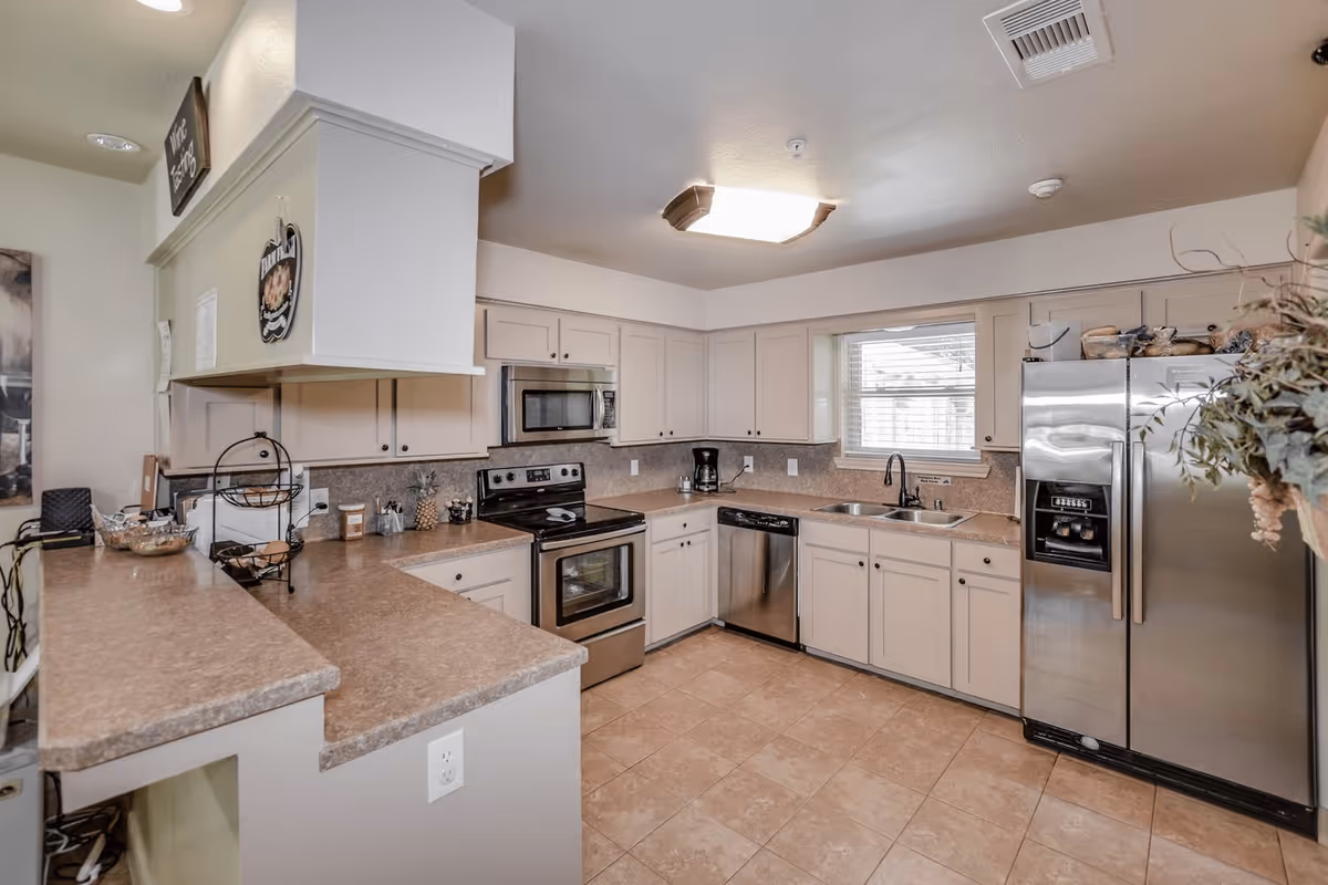 A clean and modern kitchen with beige cabinets and countertops, stainless steel appliances including a refrigerator, oven, microwave, and dishwasher. There is a double sink under a window with blinds, a coffee maker on the counter, and various kitchen items and decorations on the counters and shelves.