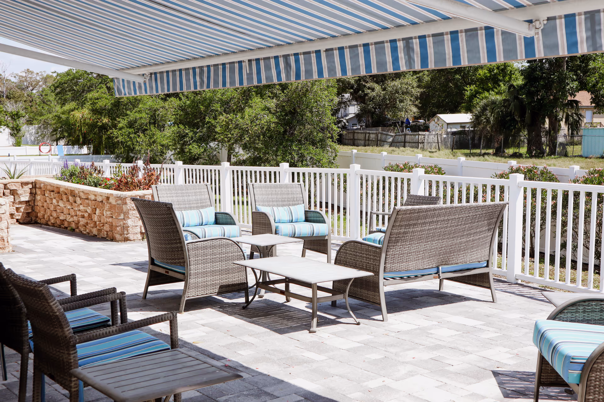 Outdoor patio area with wicker furniture including chairs and a loveseat with blue and white striped cushions, a small table, and a striped retractable awning overhead. The patio is paved with stone tiles and surrounded by a white fence with greenery and trees in the background.
